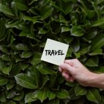 A hand holds a note with the word 'travel' against lush green leaves, symbolizing exploration.